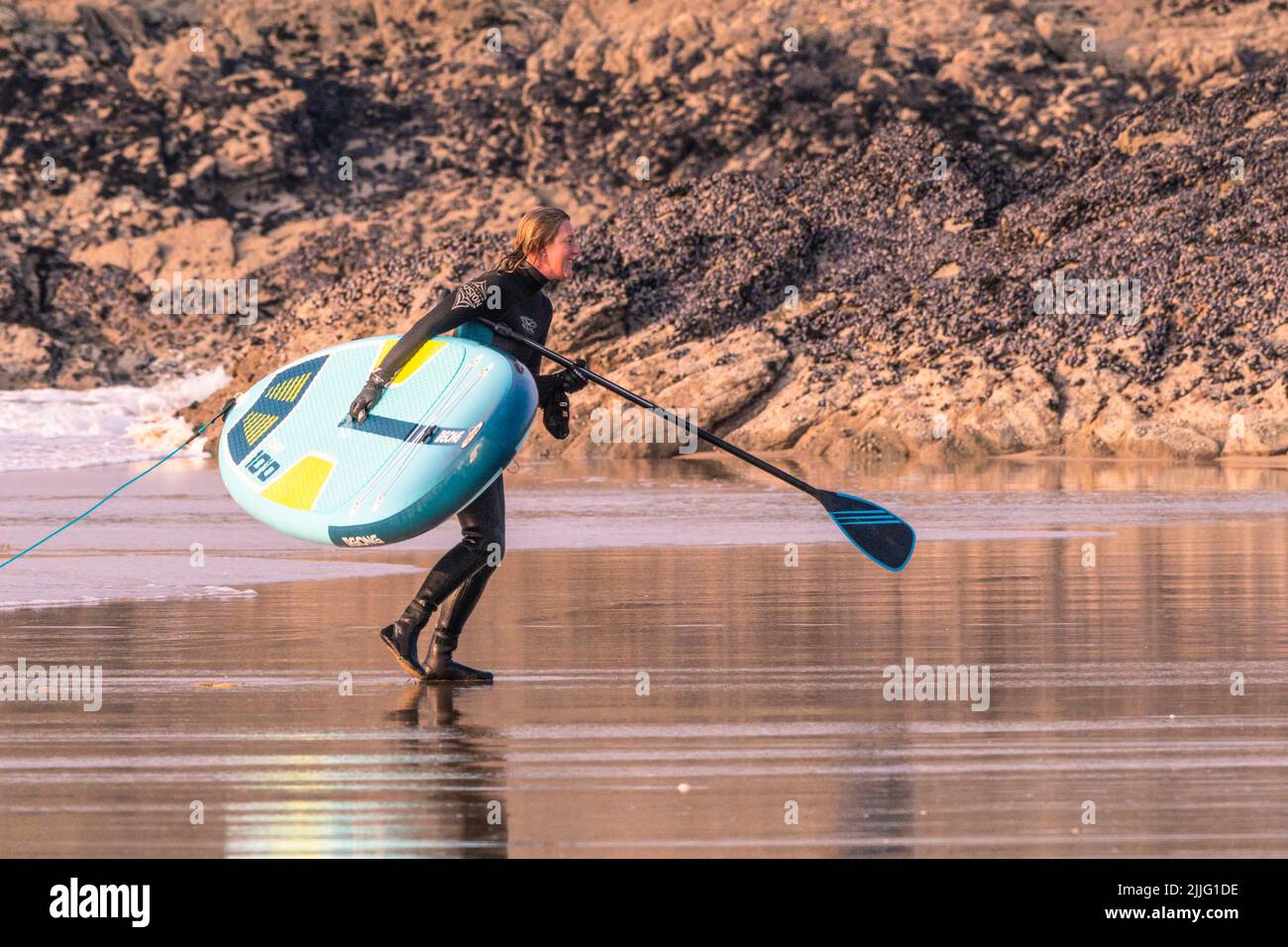 A Stand Up Paddle boarder carrying a Gong Stand Up Paddle board on Fistral Beach in evening light in Newquay in Cornwall in the UK. Stock Photo