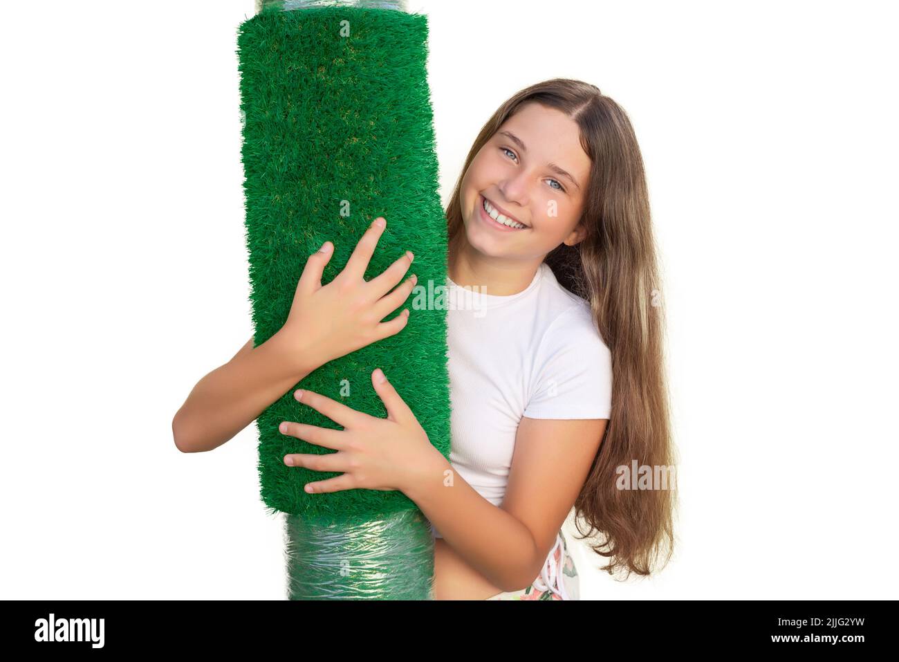 Young smiling Caucasian girl holding a roll of artificial grass on white background. Soft touch of an artificial turf sheet. Stock Photo