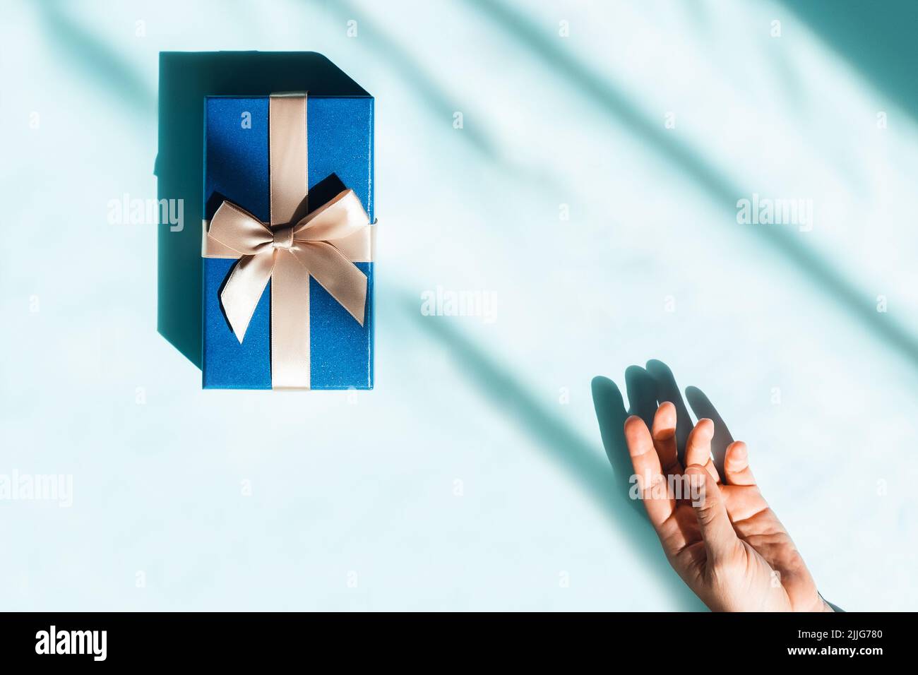 Holiday card with gift box in sunlight and women hand on blue background. Space for text. Stock Photo
