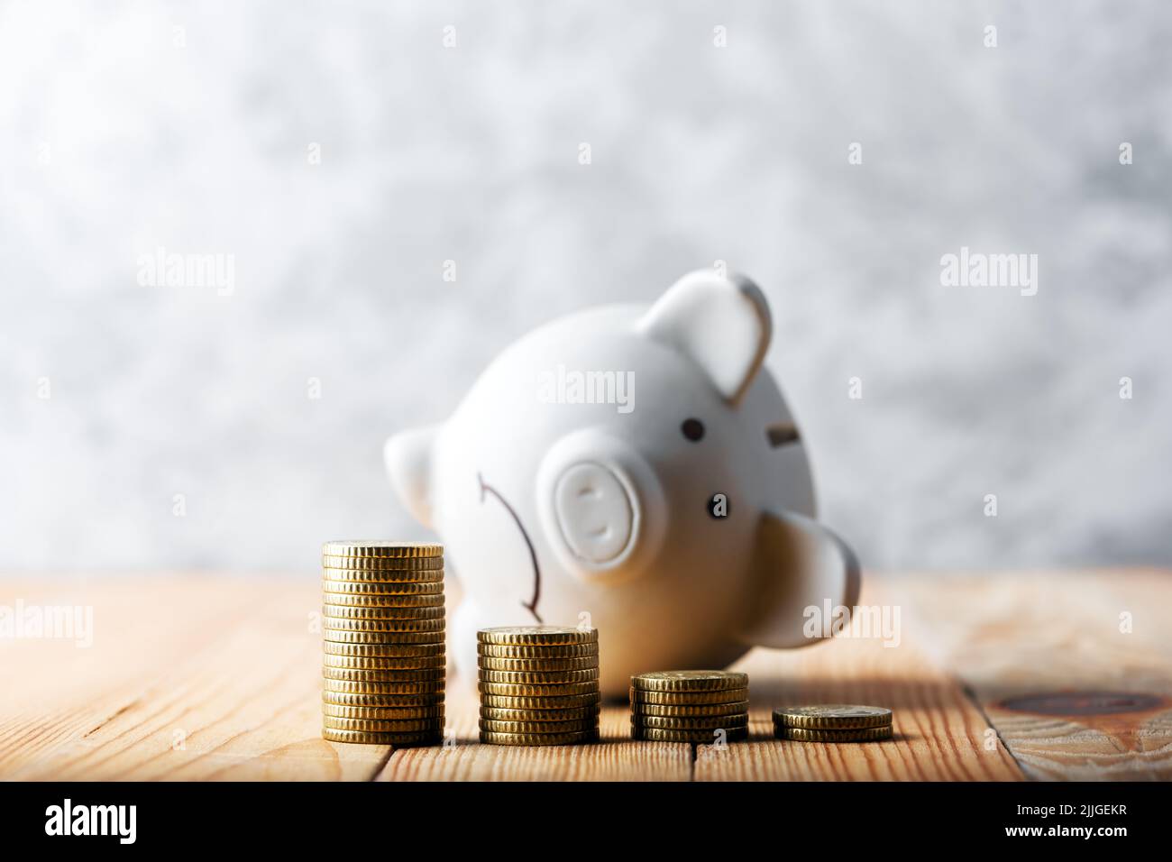 A sad piggy bank behind stacks of euro coins symbolizing the fall of monetary assets. White moneybox with coins graph. Crisis concept Stock Photo