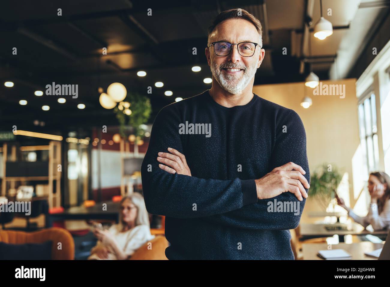 Mature businessman standing in a co-working space. Happy mature businessman smiling at the camera while standing with his arms crossed. Experienced en Stock Photo