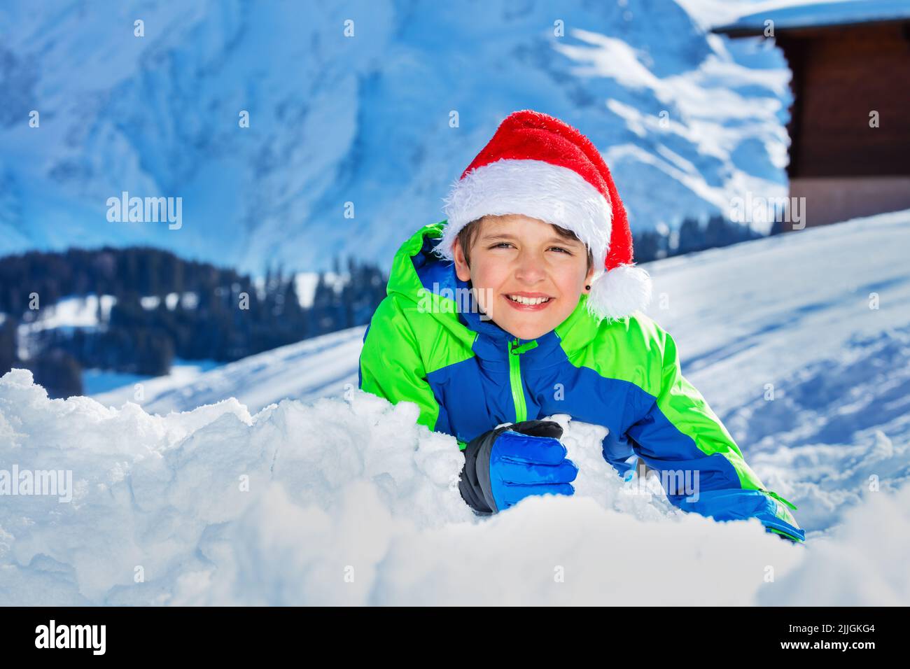 Boy with Santa hat and big smile lay in snow over mountains Stock Photo