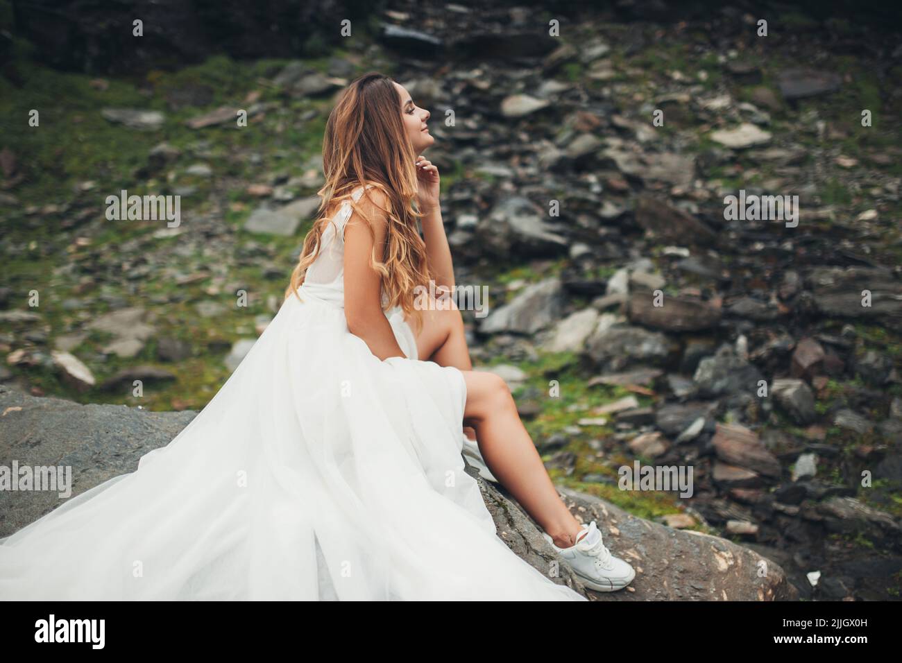 Bride in a white dress posing, sitting on a stone against backdrop of ...