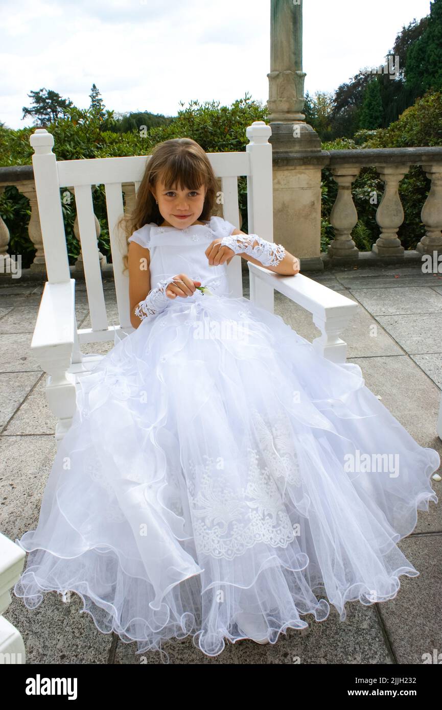 A little beautiful girl, in a white elegant dress and gloves, with flowing hair, sits on a chair.  Stock Photo