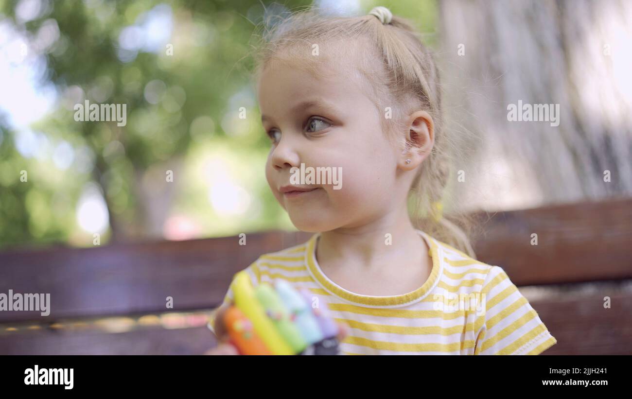 Little girl holds multi-colored gingerbread. Close-up portrait of cute child girl sitting on park bench with cookies in looks around. Stock Photo