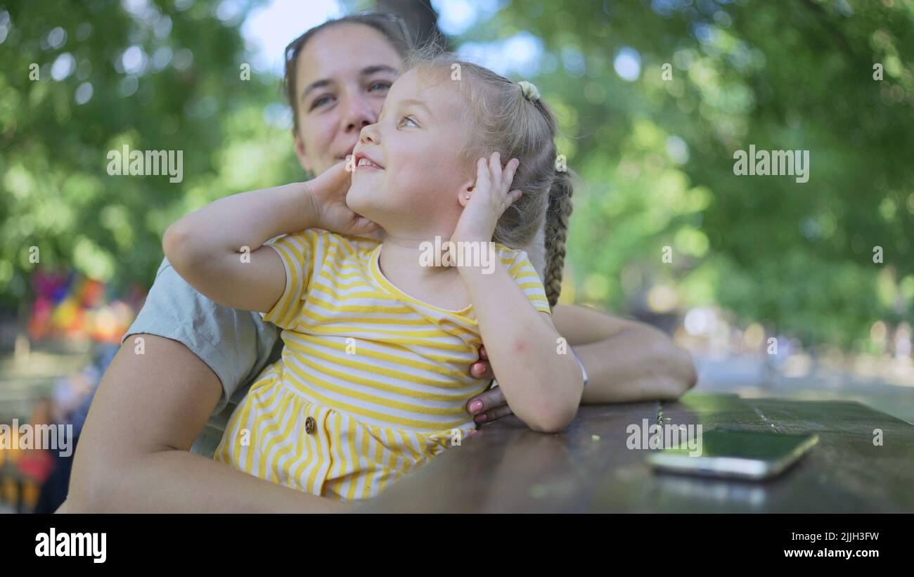 Closeup of cute little girl plays with mom while sitting in the park. Close-up of mother and daughter playing while sitting on a park bench in a stree Stock Photo