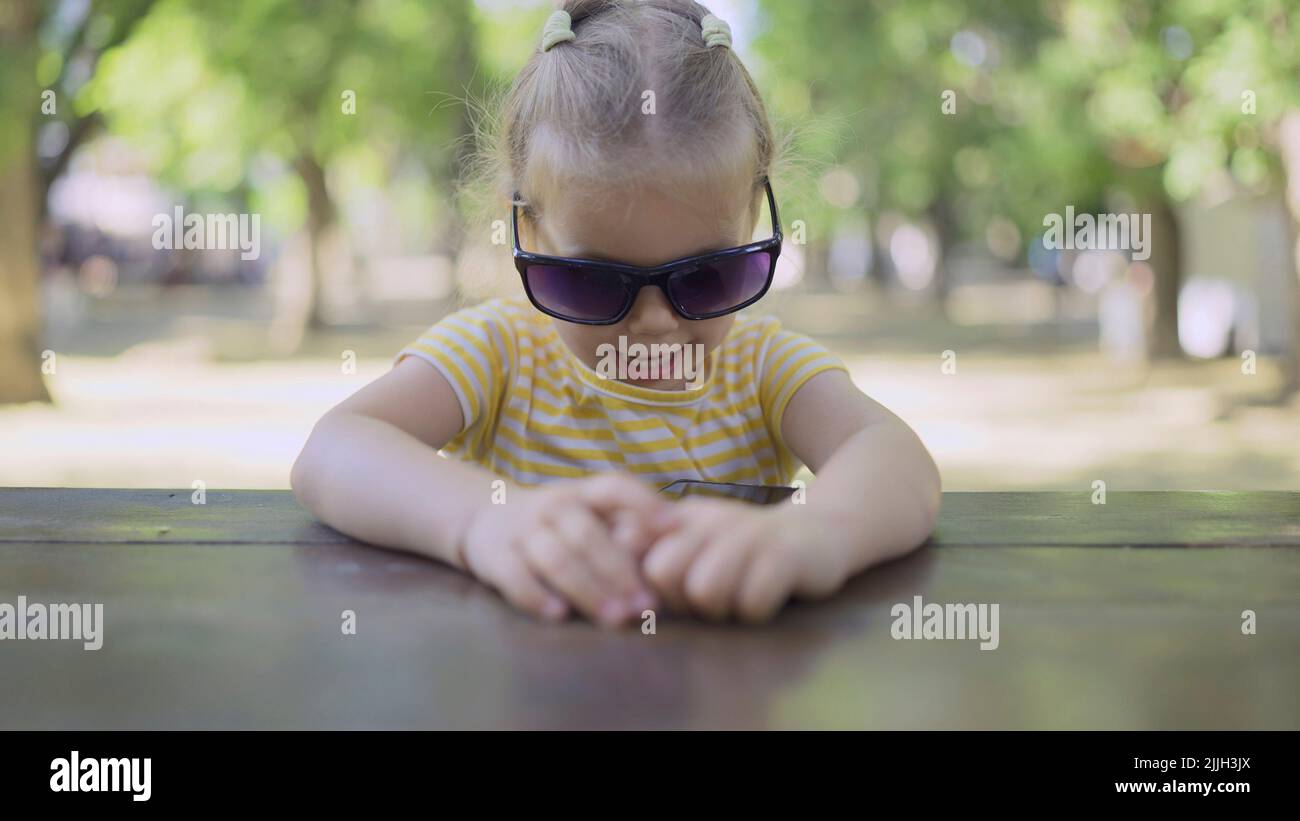 Little girl in mom's sunglasses is learning foreign language by repeating words from mobile phone. Close-up portrait of child girl sitting in city par Stock Photo