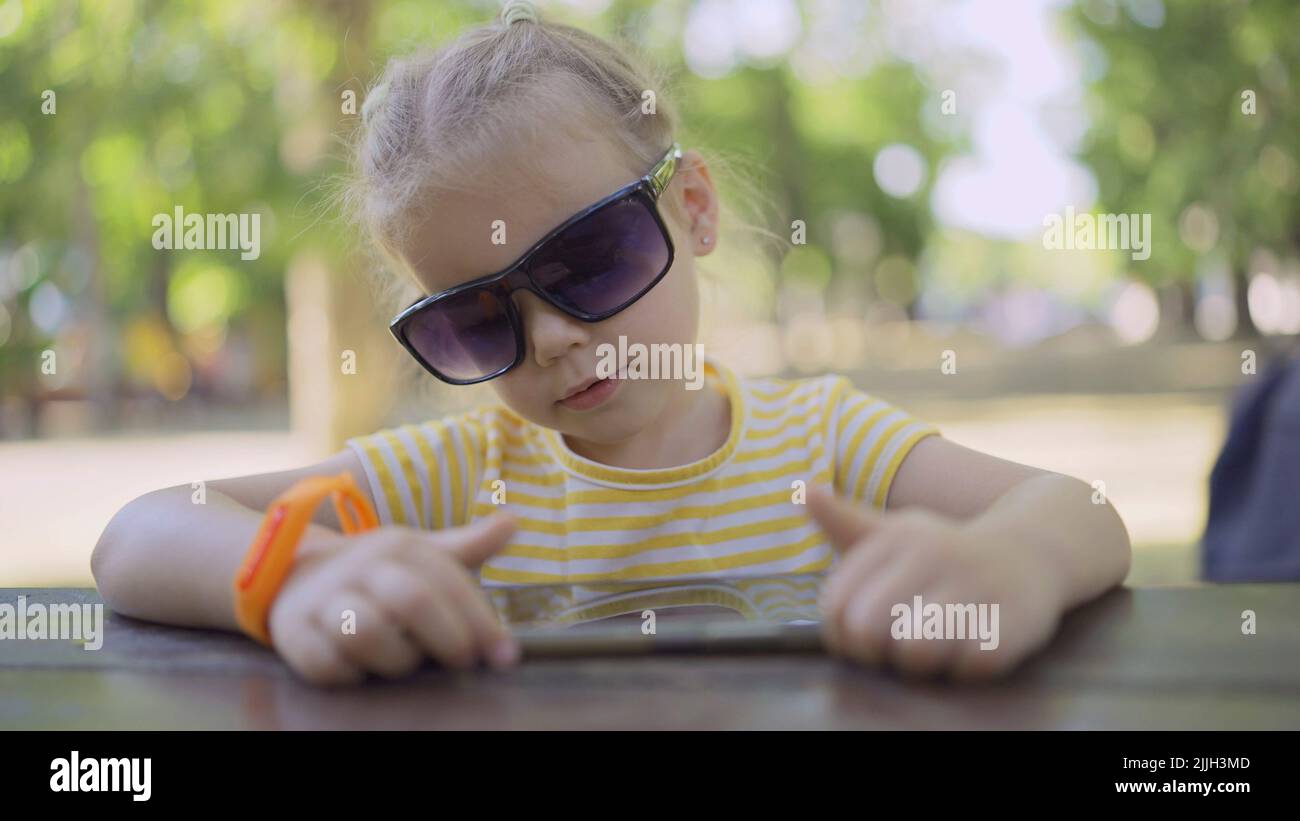 Little girl in mom's sunglasses is learning foreign language by repeating words from mobile phone. Close-up portrait of child girl sitting in city par Stock Photo