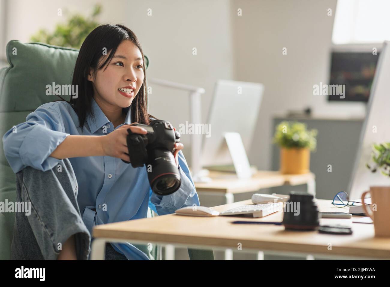 Cheerful Vietnamese Woman Photographer Holding Photocamera Sitting At Workplace Stock Photo