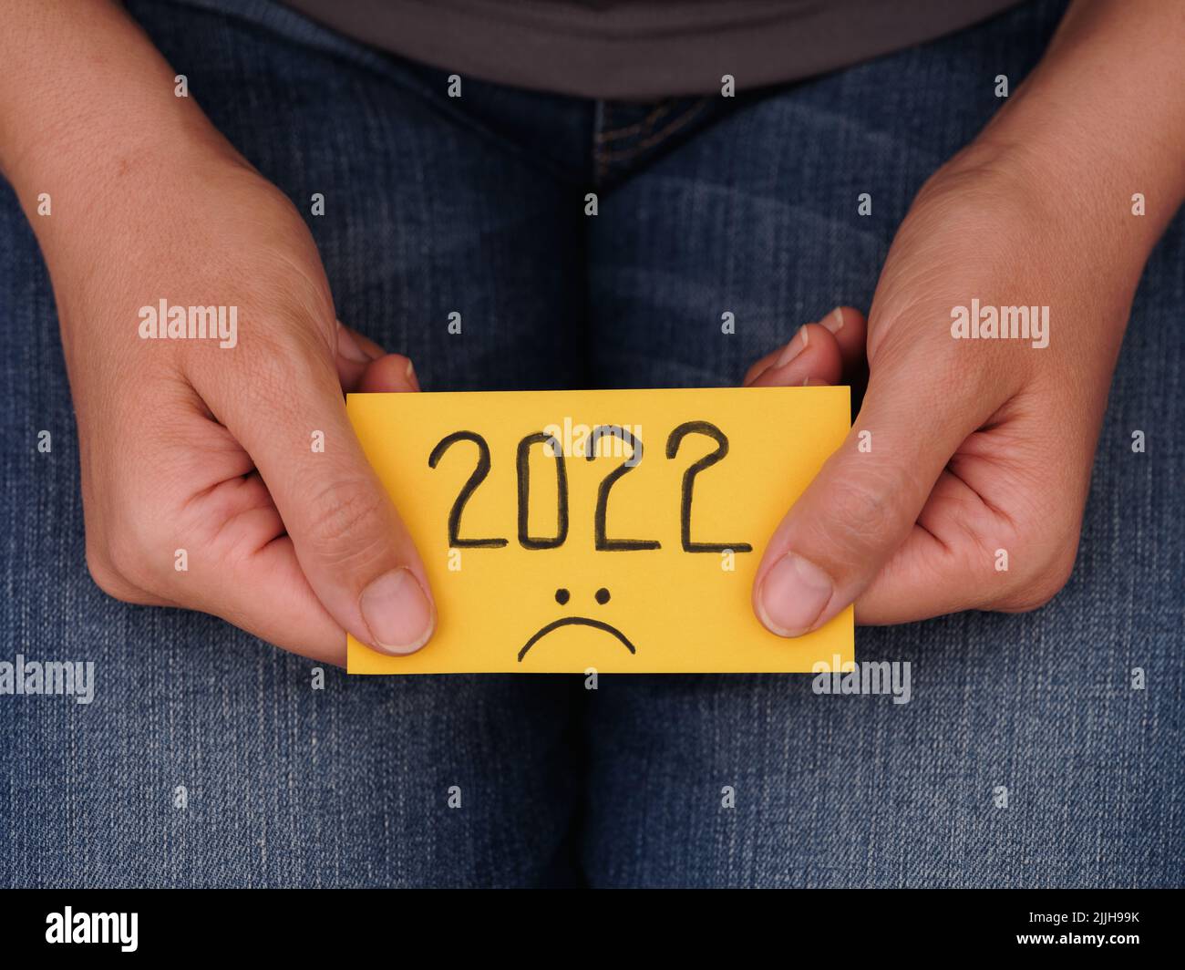 A woman holding a piece of yellow paper with the year 2022 and a sad face on it in her hands. 2022 horrible year concept. Close up. Stock Photo