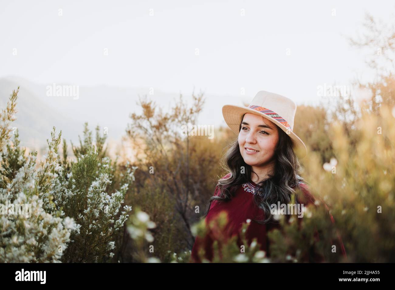 Smiling latin backpacker woman wearing a traditional red poncho and a hat in a leafy natural space at sunset Stock Photo