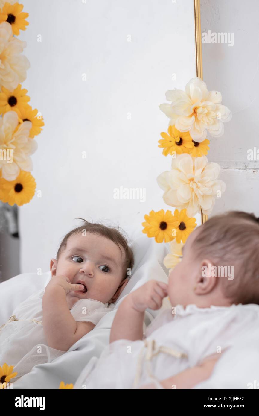 beautiful latina baby, impressed while looking at her reflection in the mirror adorned with yellow and white flowers, with one hand touching her mouth Stock Photo