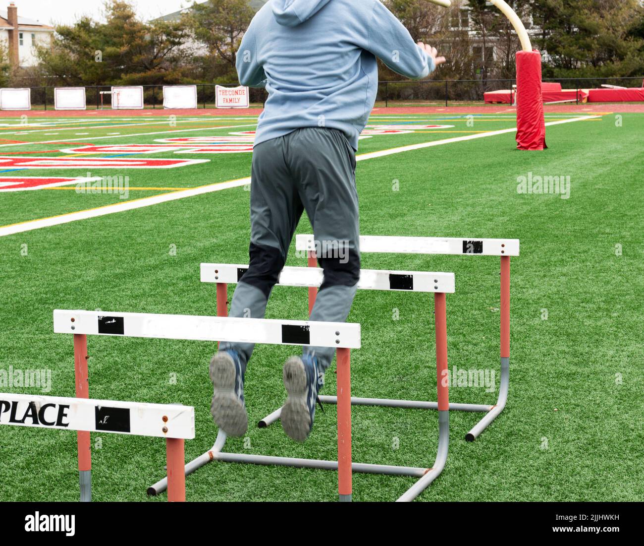 Rear view of a high school boy is jumping over track hurdles on a turf