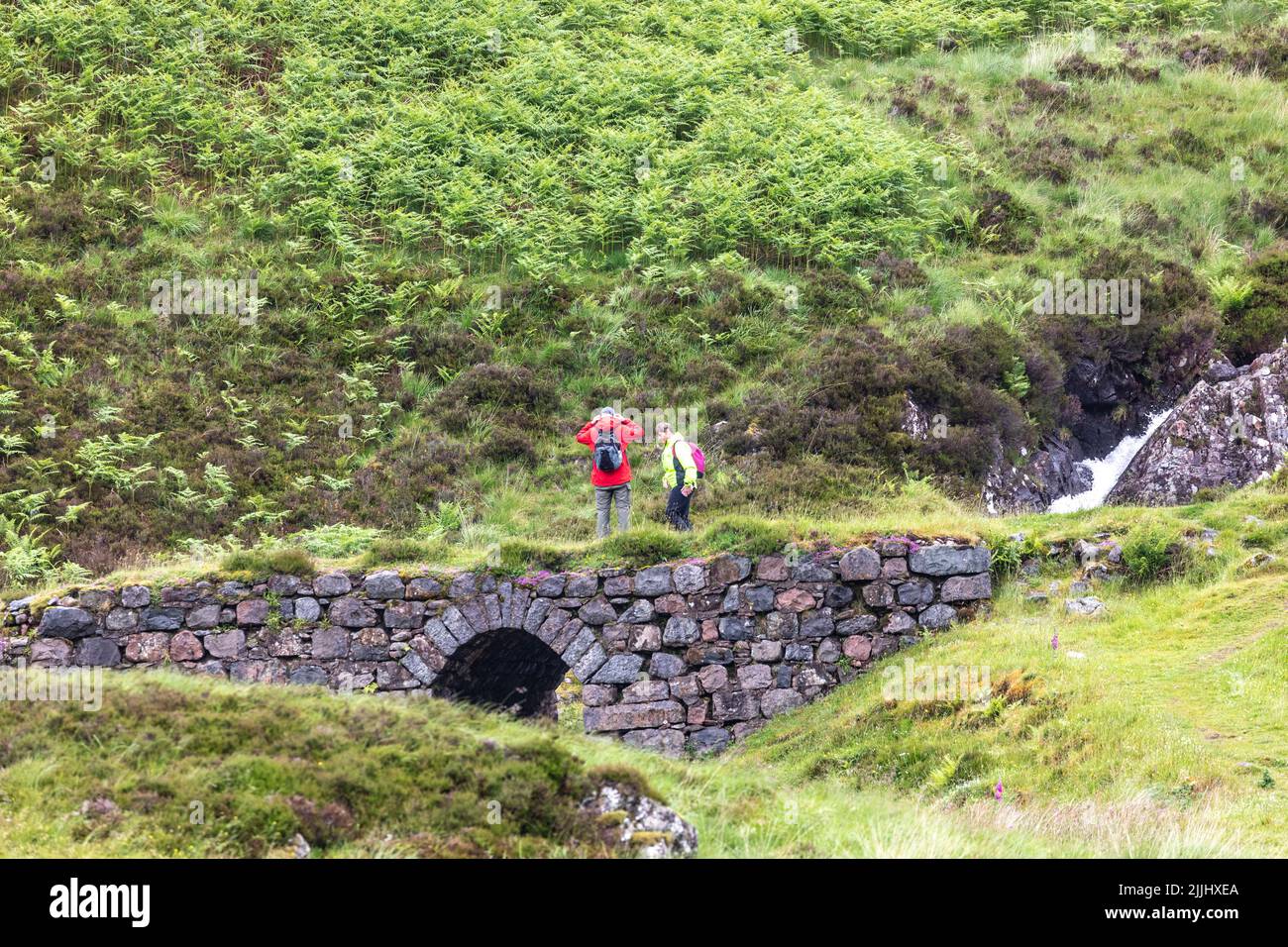 Couple hiking in Glen Coe stop beside a waterfall and small stone bridge, Scottish highlands,Scotland,UK summer 2022 Stock Photo