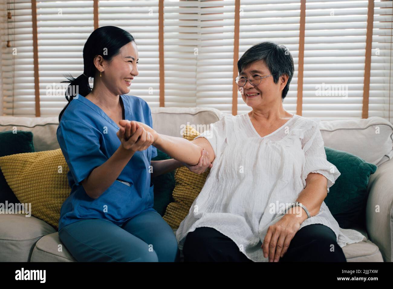Delightful Female Caregiver In Blue Uniform Smiling And Stretching Arm Of Elderly Asian Woman