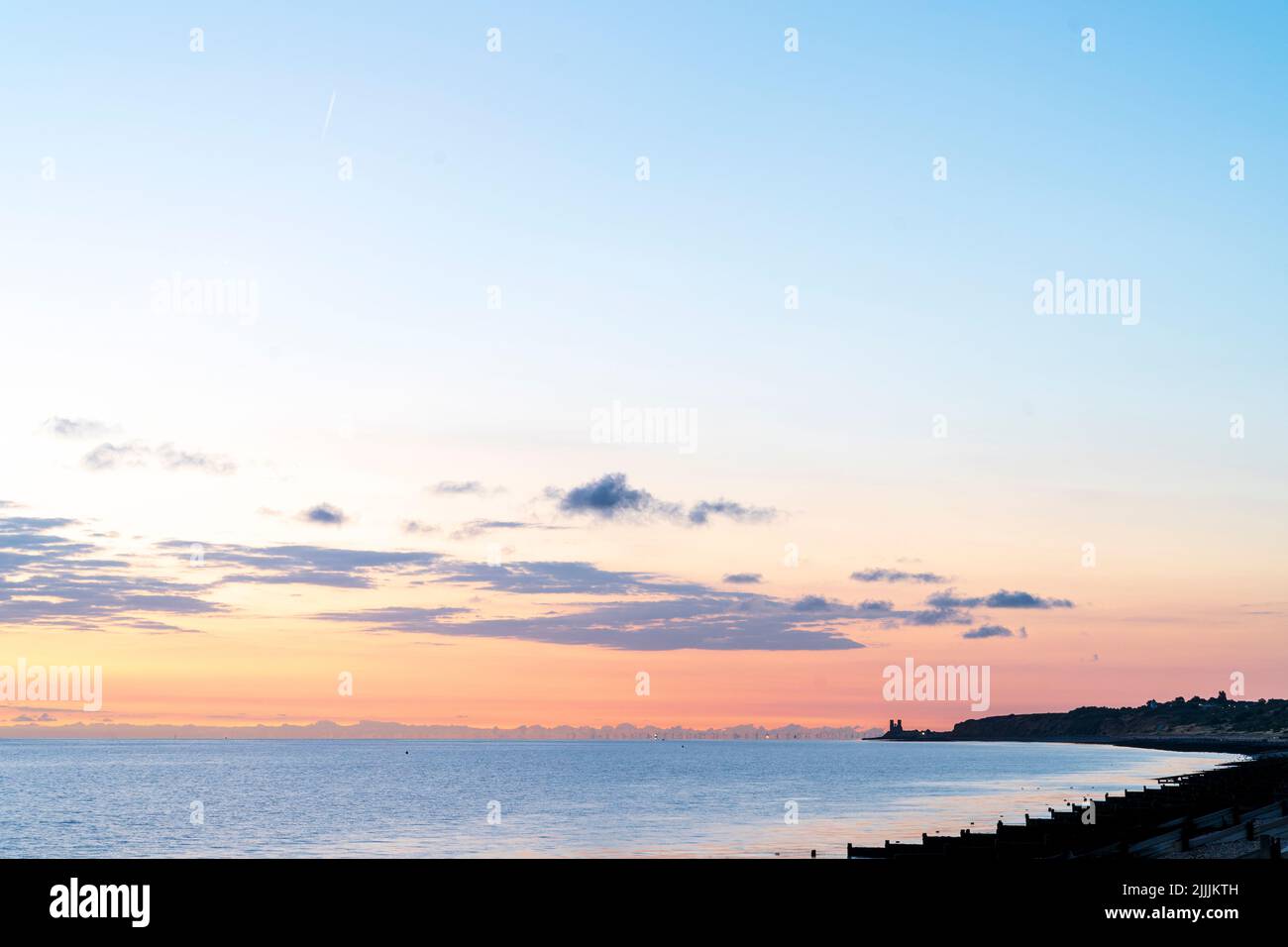 The dawn sky over the North Sea seen from the Kent coast at Herne Bay. Silhouette of some breakwaters, groynes in the foreground and the wide bay ending at the landmark twin towers of the ruins of Reculver church in the distance. Some scattered clouds in the sky. Stock Photo