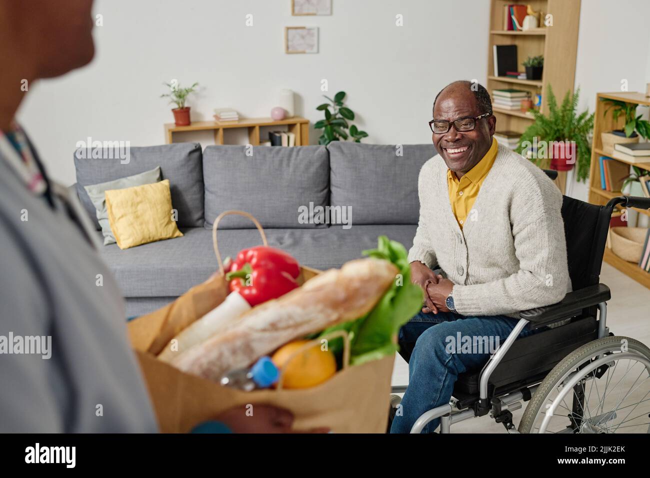 African happy man with disability sitting in wheelchair and meeting caregiver bringing groceries for him Stock Photo