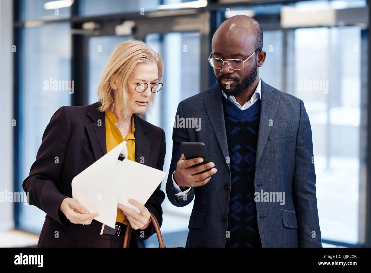 Mature female lawyer with documents discussing the deal with her colleague while he using his mobile phone before the meeting Stock Photo