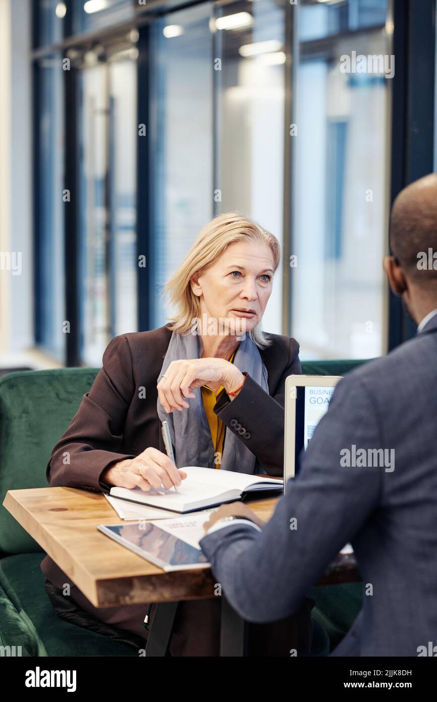 Serious mature businesswoman listening to her partner during business meeting while they sitting at the table with documents and laptop Stock Photo