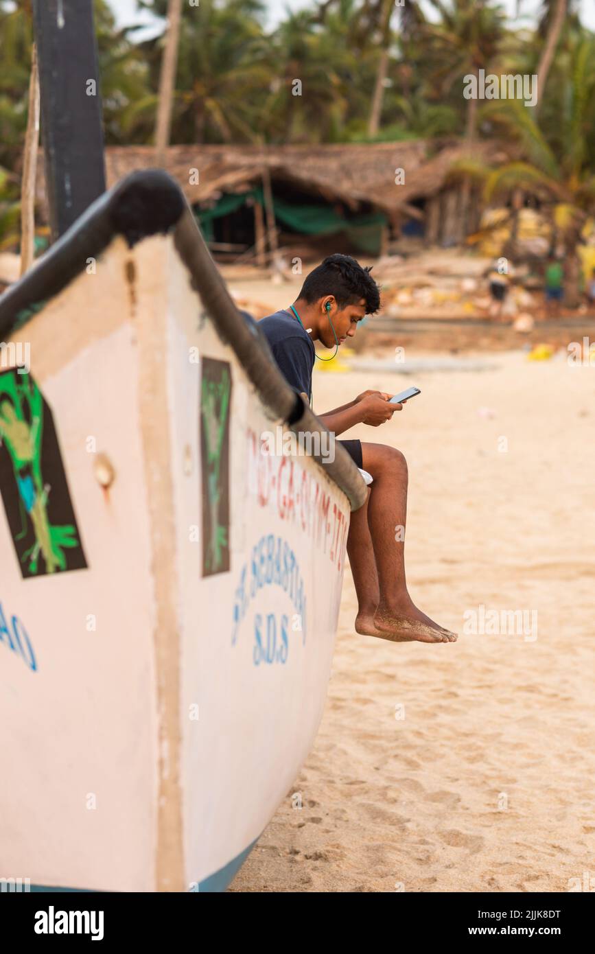 A young Indian man sitting on the boat and listening to music at Ashwem Beach in Goa India Stock Photo
