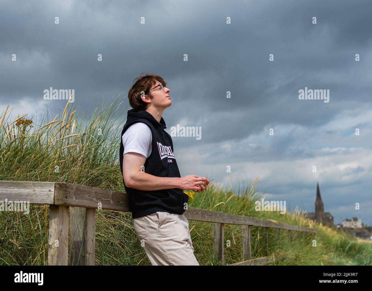 Natural portrait of a young man standing on a fence on the boardwalk at Longsands beach in Tynemouth with the church in the background Stock Photo