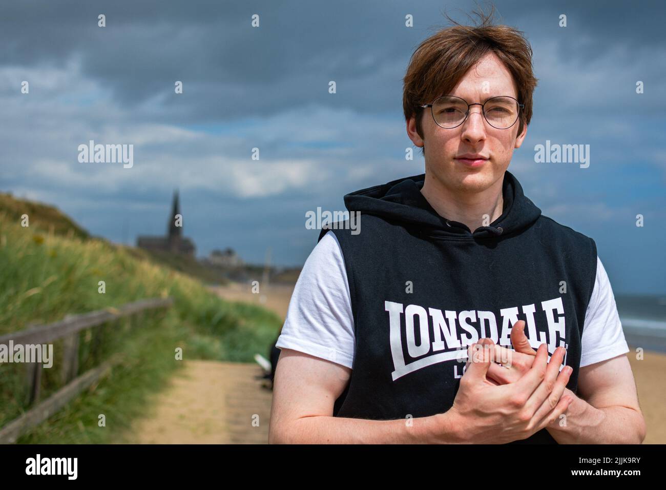 Natural half portrait of a young man wearing sleeveless hoodie and white t-shirt at the beach in Tynemouth clasping hands Stock Photo