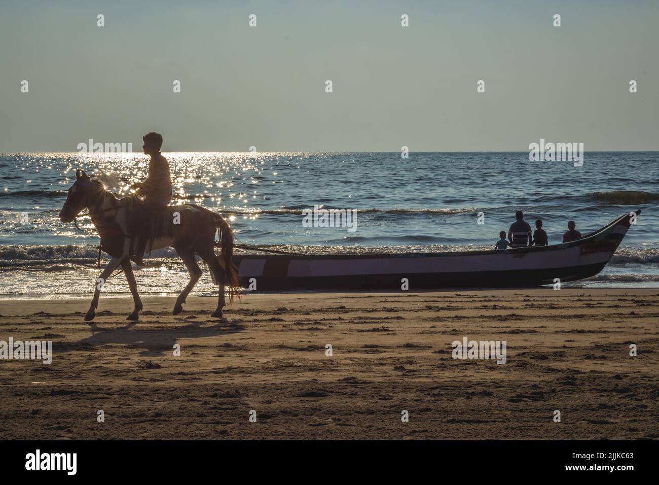 A young male riding a horse and a boat with a family on the sunny shore in Chennai, India Stock Photo