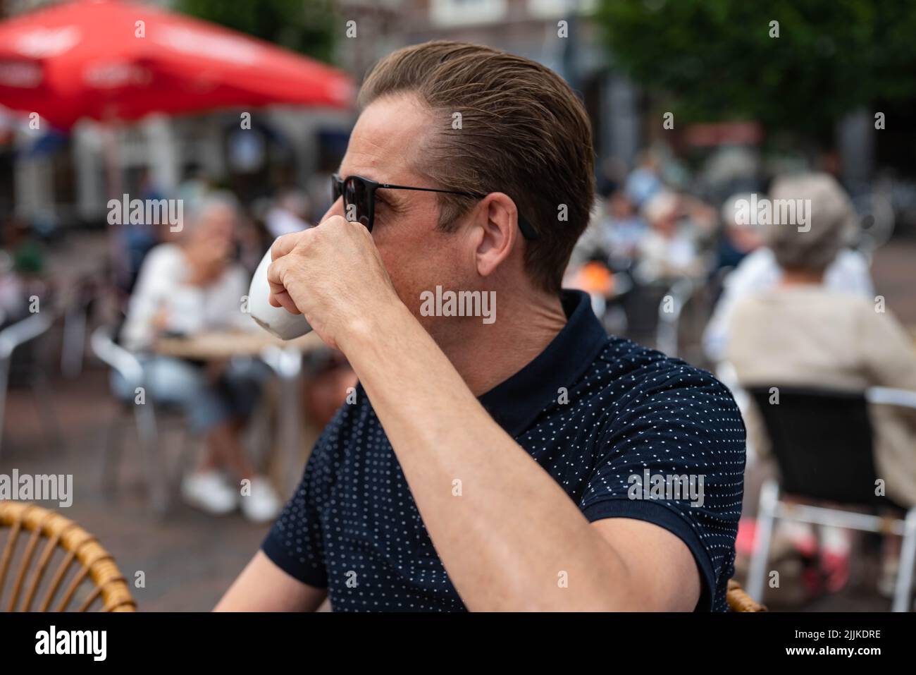 White Caucasian man with sunglasses looks past the camera and drinks coffee from a cup on a terrace during the summer. Stock Photo