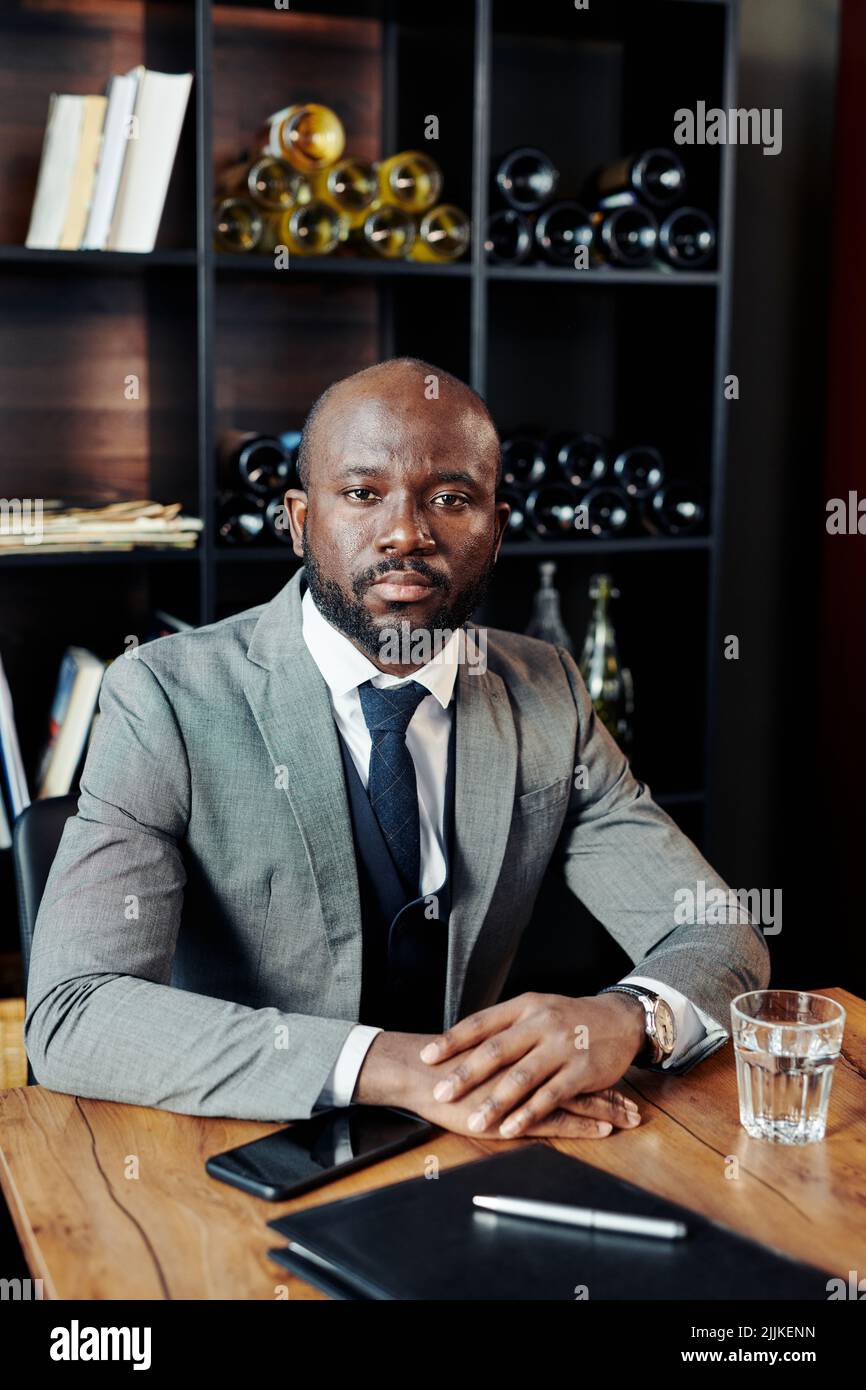 Portrait of serious African businessman in suit sitting at the table and looking at camera while waiting for somebody at restaurant. Stock Photo