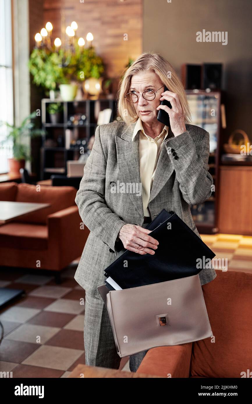 Serious mature businesswoman talking on mobile phone with her client while putting the folder in her bag after meeting at the restaurant Stock Photo