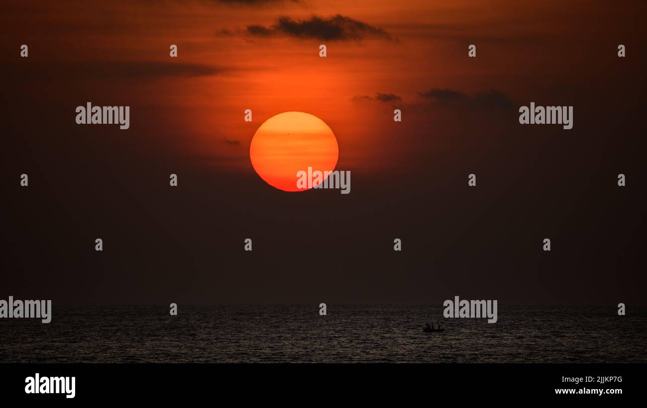 Big ball of the orange sun setting behind the ocean horizon, beautiful sunset colors in the sky. Solitary silhouette fishing boat sails in the ocean. Stock Photo