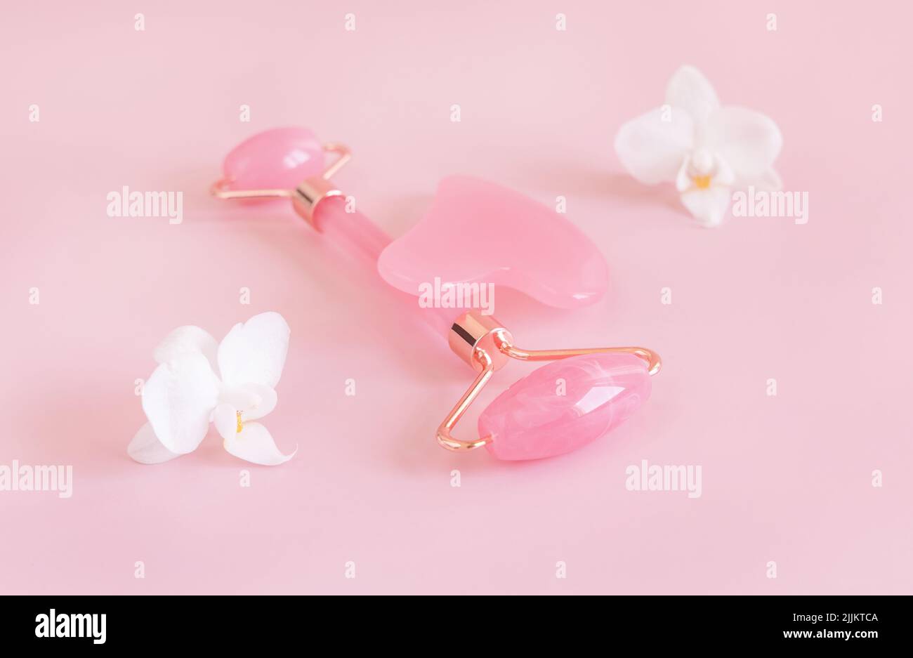 Rose quartz facial roller and gua sha massager near white orchid flowers, close up on light pink. Anti-age massage and natural lifting therapy at home Stock Photo
