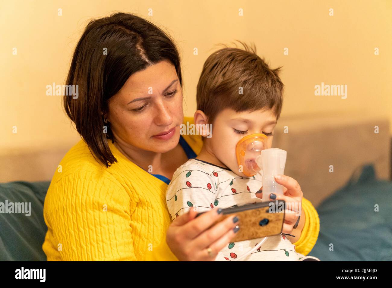A closeup of a woman giving a child inhalation while he is watching something on a smartphone. Stock Photo
