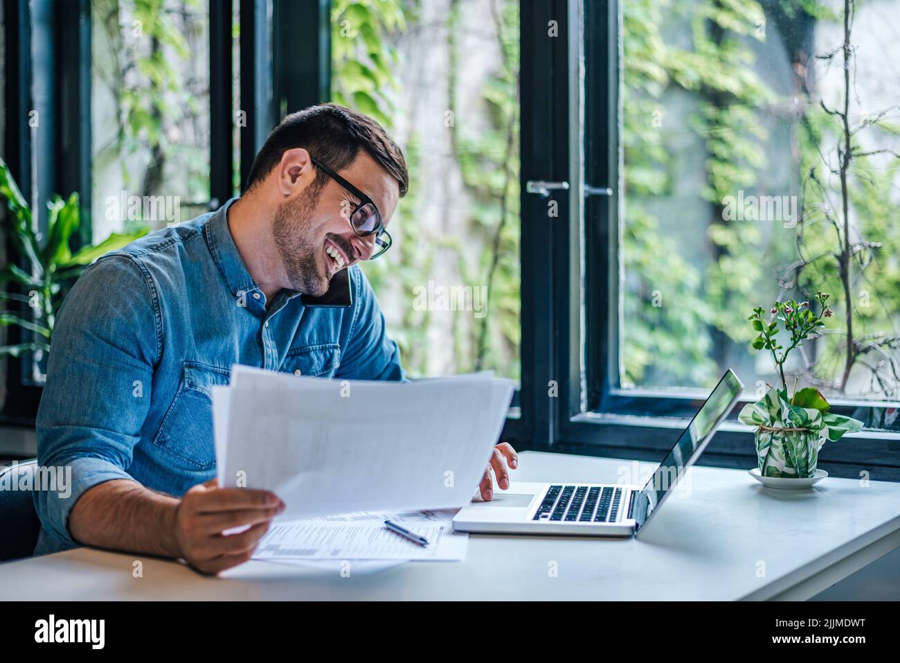 Happy male professional discussing on smart phone while using laptop. Male entrepreneur with documents is multi tasking at table. He is wearing casual Stock Photo