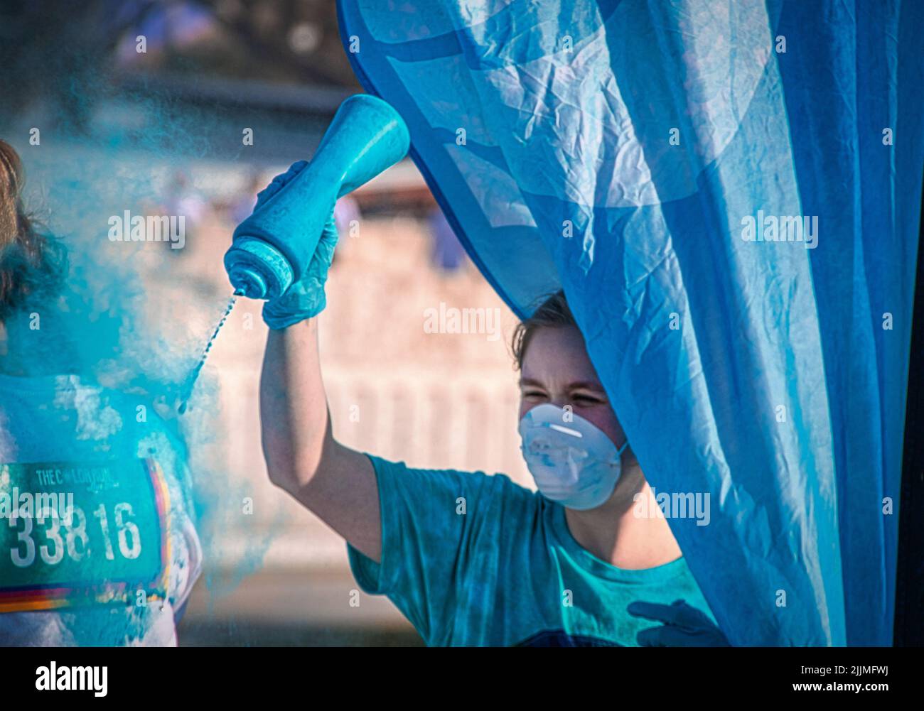 April 6 2019 Tulsa USA - Holi - Color Run in Tulsa USA - Volunteer in mask  standing behind flag squirts turquoise powder on runner against blurred ba Stock Photo