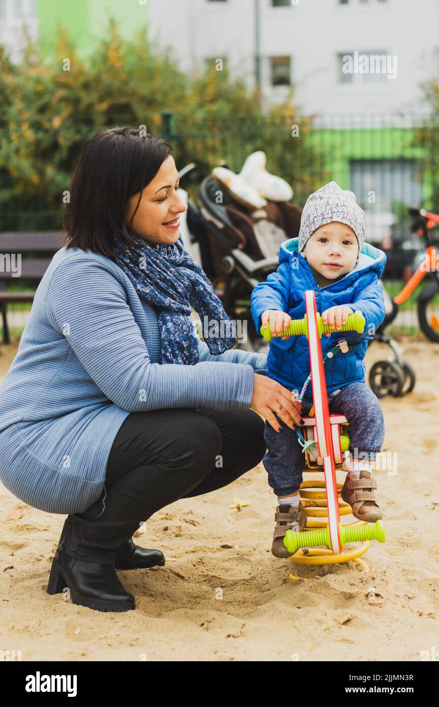 A Caucasian mother next to a toddler sitting on a swing at a playground in the Orla Bialego district, Poznan, Poland Stock Photo