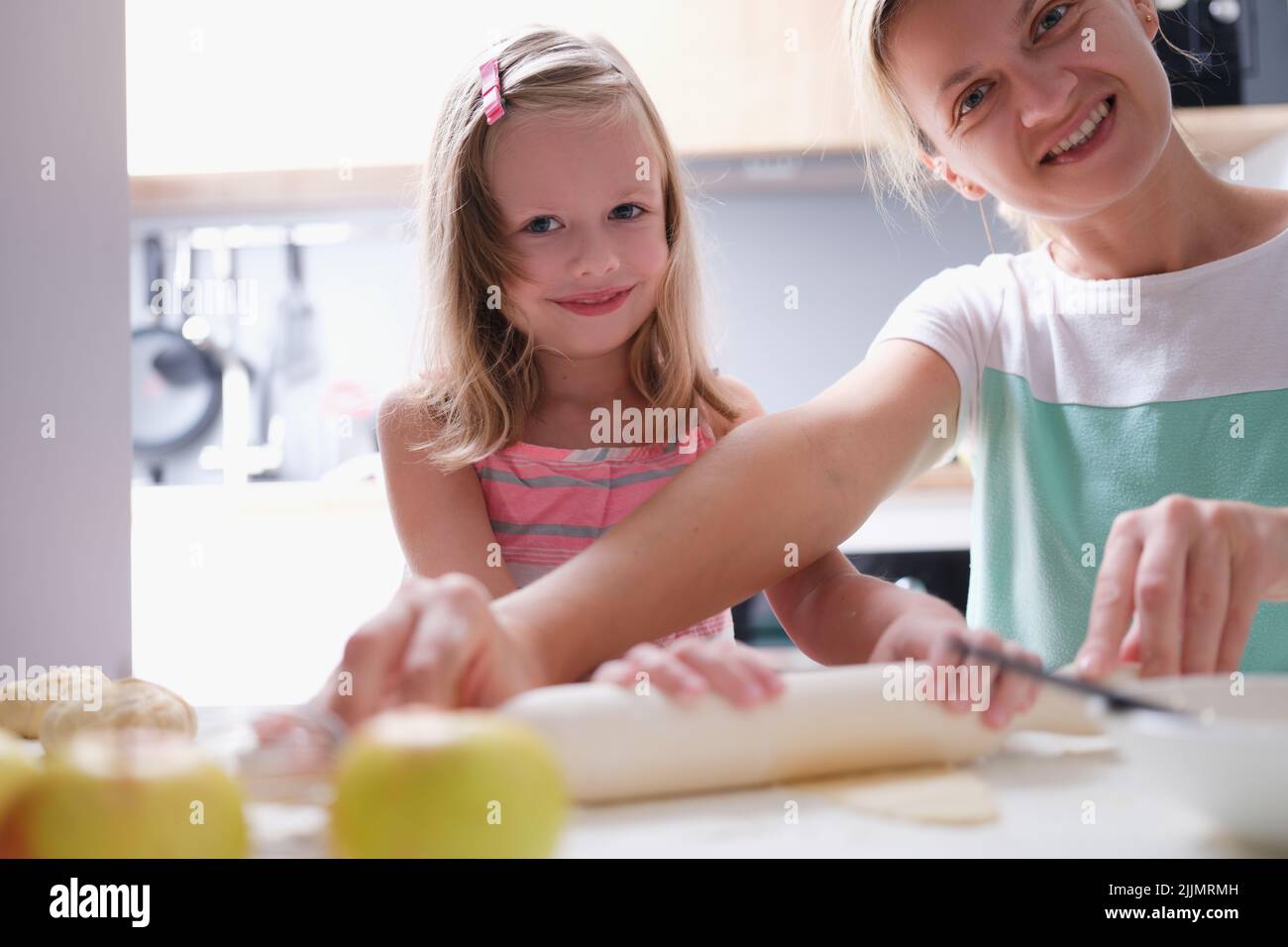 Happy young mother and little child daughter rolls out dough on kitchen table with pin Stock Photo