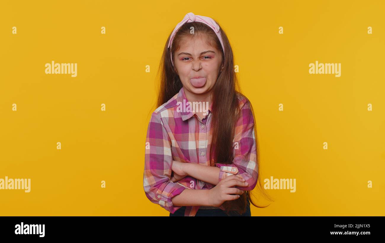 Cheerful funny young preteen child girl kid showing tongue making faces at camera, fooling around, joking, aping with silly face, teasing. Little toddler children isolated on studio yellow background Stock Photo