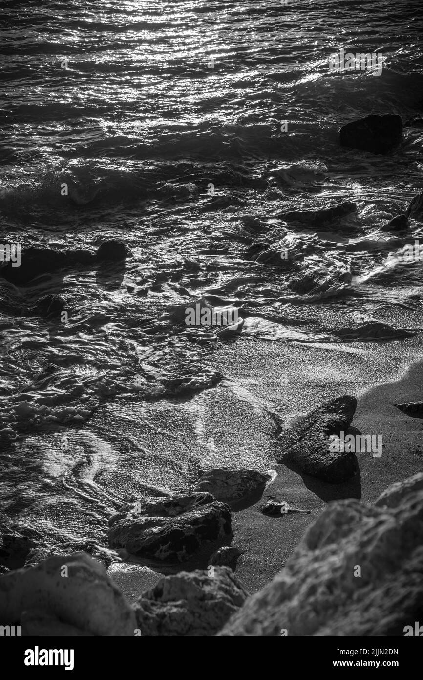 A grayscale shot of waves hitting the sandy beach during sunset Stock Photo