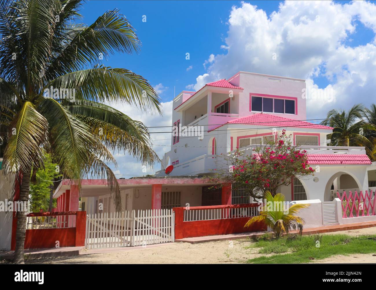 Cute pink and white Mexican three story house with red fence and palm and flowering trees against a beautiful blue sky with fluffy clouds Stock Photo