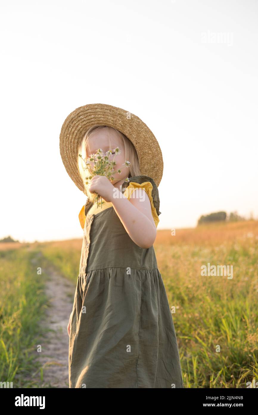 A little blonde girl in a straw hat walks in a field with a bouquet of ...