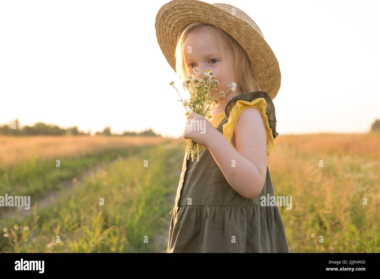 A little blonde girl in a straw hat walks in a field with a bouquet of ...
