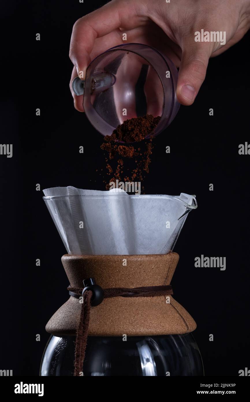 A vertical closeup of the hand adding fresh coffee grinds into a glass coffeemaker. Stock Photo