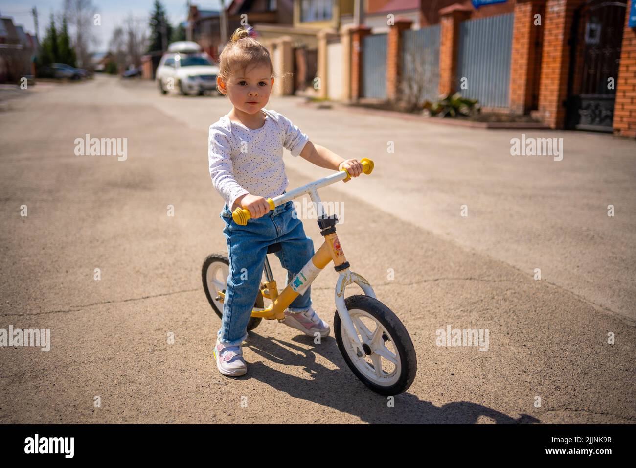 Cute little toddler girl in blue overalls riding on run balance bike. Happy healthy lovely baby child having fun with learning on leaner bicycle Stock Photo