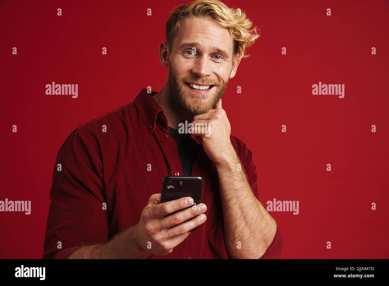 Bearded blonde man smiling and using mobile phone isolated over red background Stock Photo