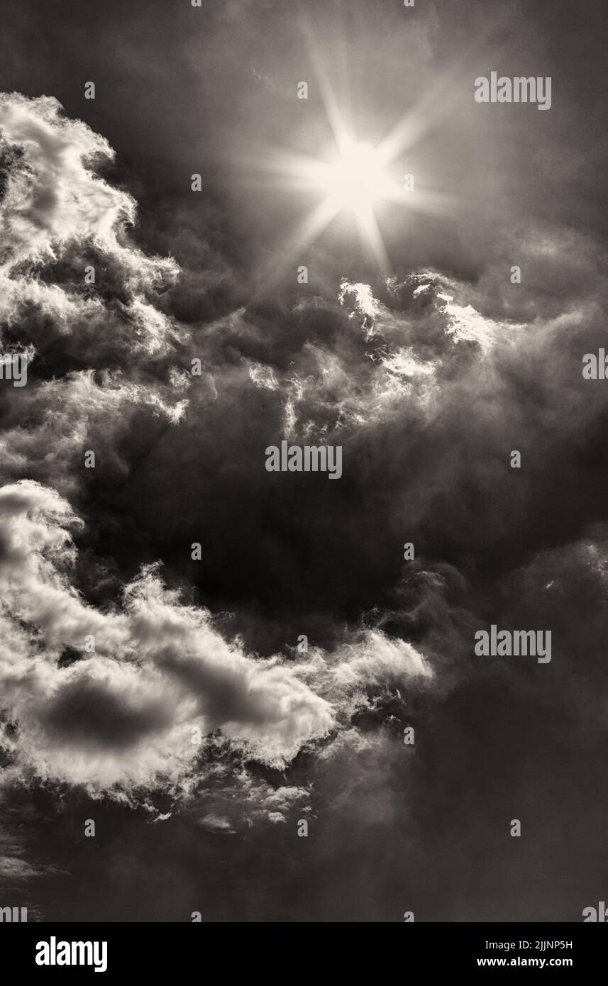 A vertical shot of a cloudy sky with a shining sun in grayscale Stock Photo