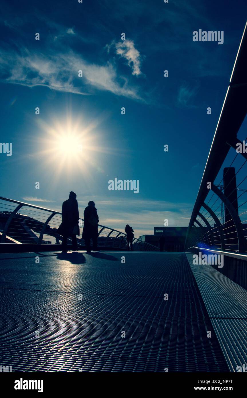 A vertical shot of peoples silhouettes on a bridge with a bright sun in ...