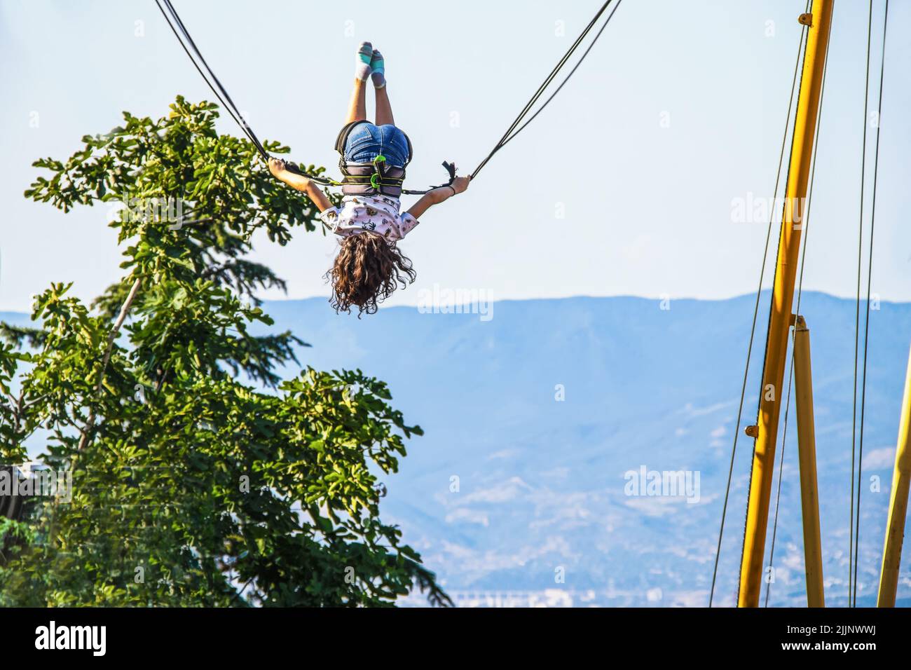 Little girl with jean shorts and long hair upside down on bouncing bungee swing overlooking vista of mountain and valley Stock Photo