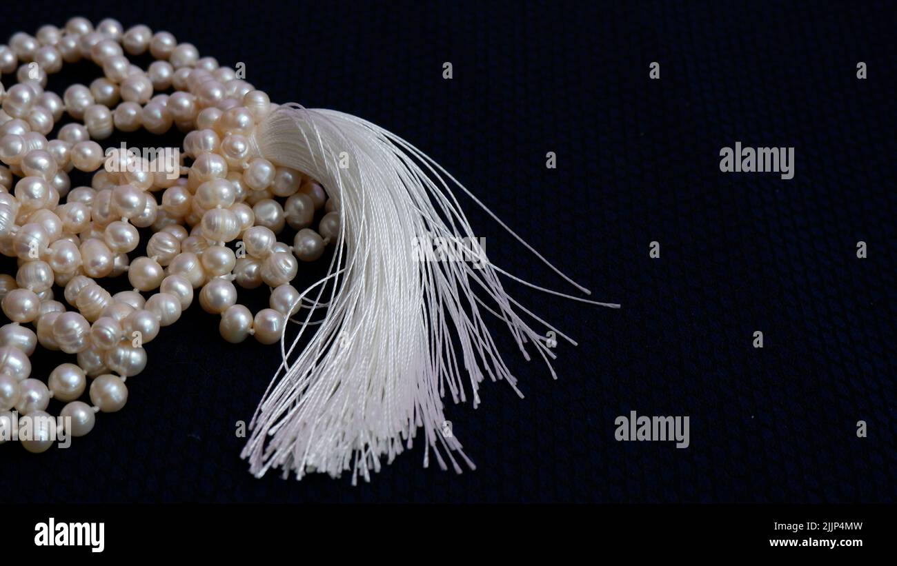 On a dark blue background lies a pearl necklace and a white silk tassel. Jewelry for the bride for the wedding. Stock Photo