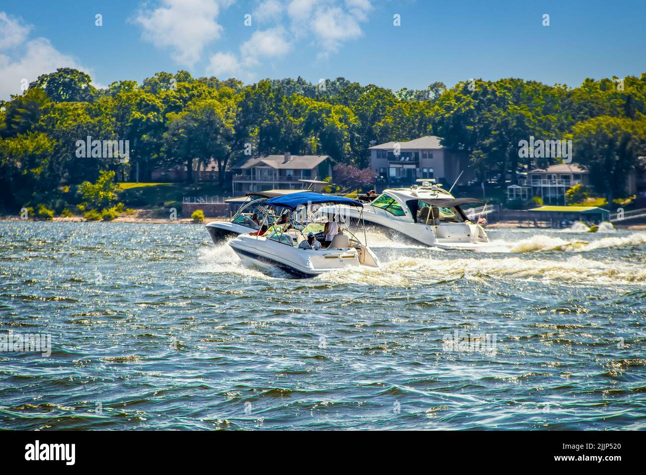 Three speedboats at lake racing in choppy water past lakeside homes and trees on nearby shore on sunny day - selective focus. Stock Photo